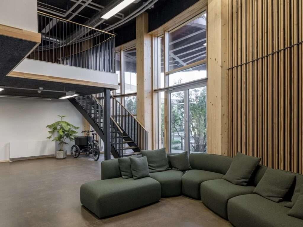 Double-height communal lounge inside TRAE tower with a large green sectional sofa, a black steel staircase, and floor-to-ceiling windows showing timber framework.