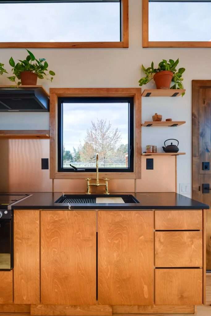 Modern tiny home kitchen with natural wood cabinetry, black countertops, a copper backsplash, a golden faucet, and an integrated window view.