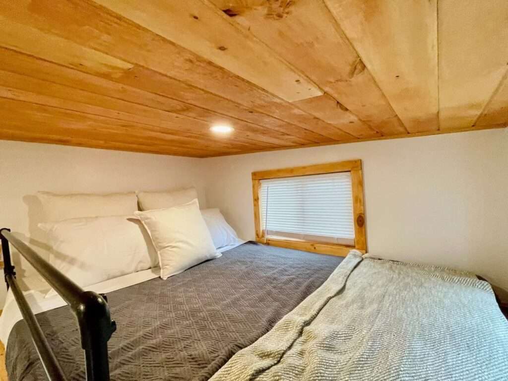 Close-up of a queen-sized bed in the loft of Rasa tiny home with natural wood ceiling, white walls, and a small side window.