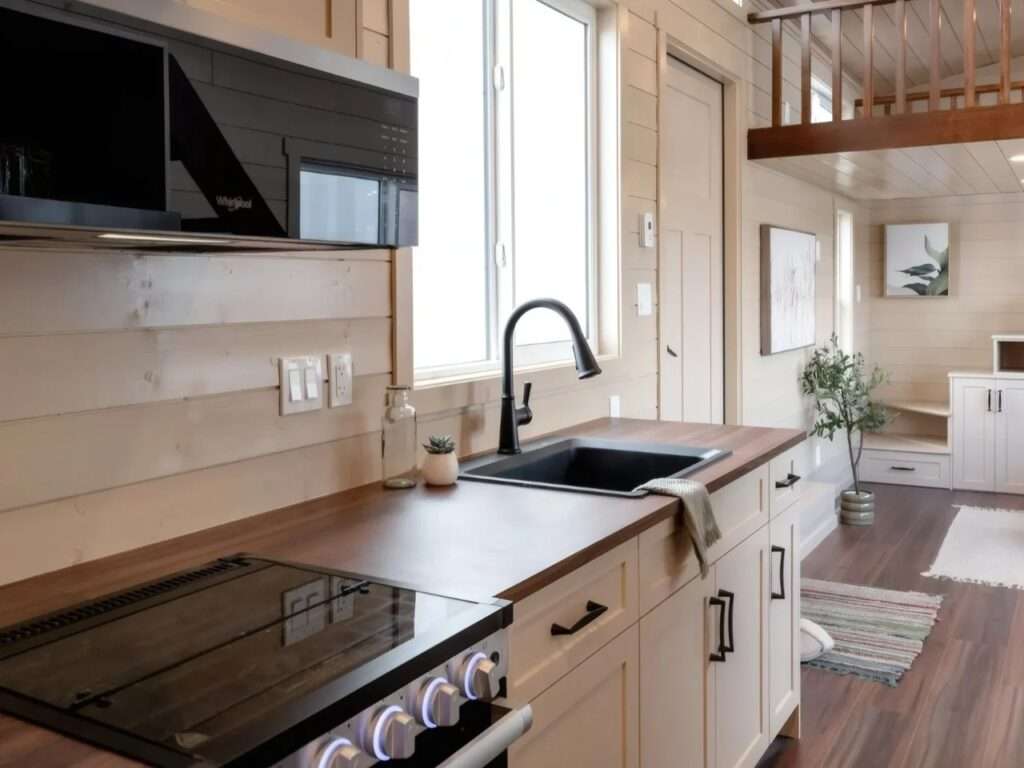 Close-up of the Ruby tiny home kitchen featuring a dark wood countertop, a black undermount sink with a gooseneck faucet, a modern cooktop, and an over-the-range microwave.