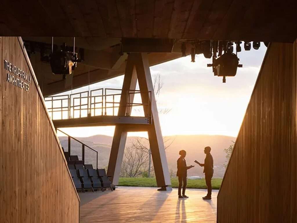 Two people conversing on the wooden stage of the open-air theater during sunset framed by a structural timber column.
