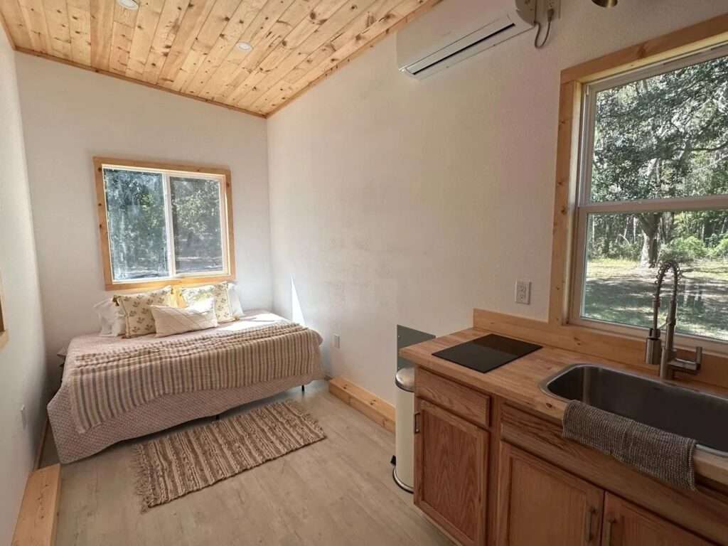 Interior shot of Shanty tiny house showing a queen-sized bed on the ground floor next to a window and a kitchenette with wooden countertops.