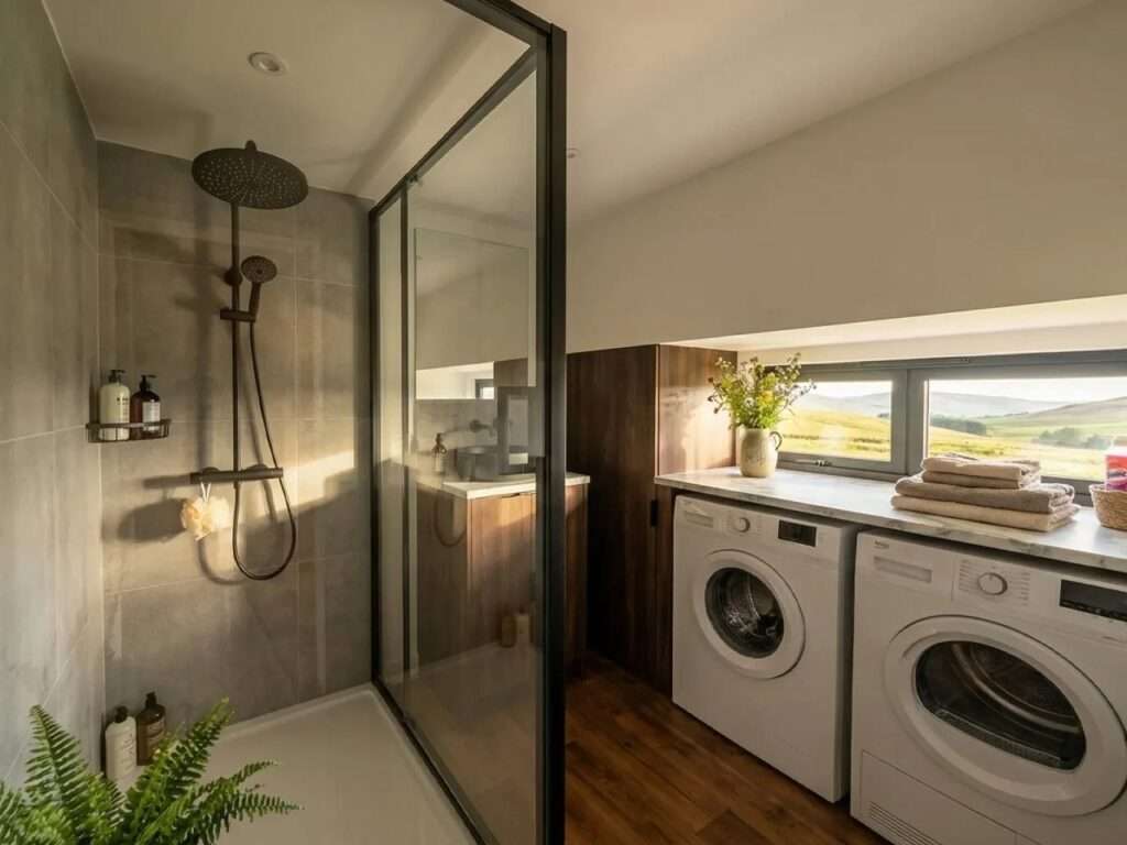 Modern tiny house bathroom with a glass walk-in shower, grey tiling, and integrated laundry area featuring a washing machine and dryer.