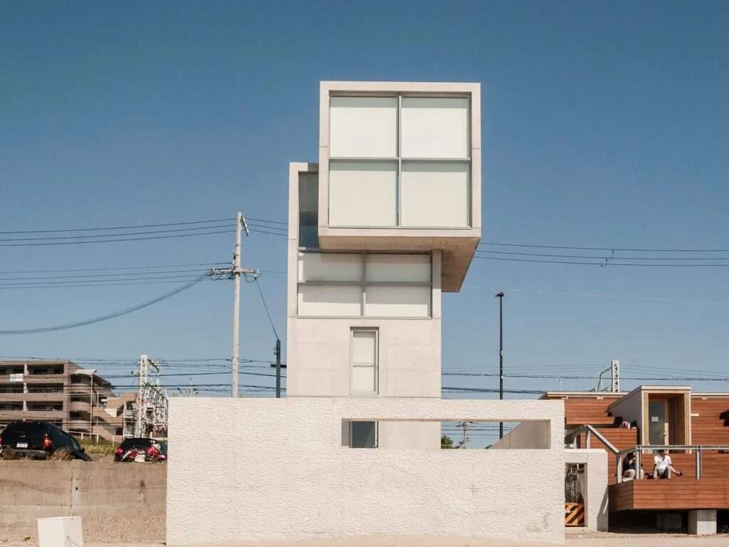 Street-level view of the concrete 4x4 House behind a textured white concrete perimeter wall, framed against a clear sky.