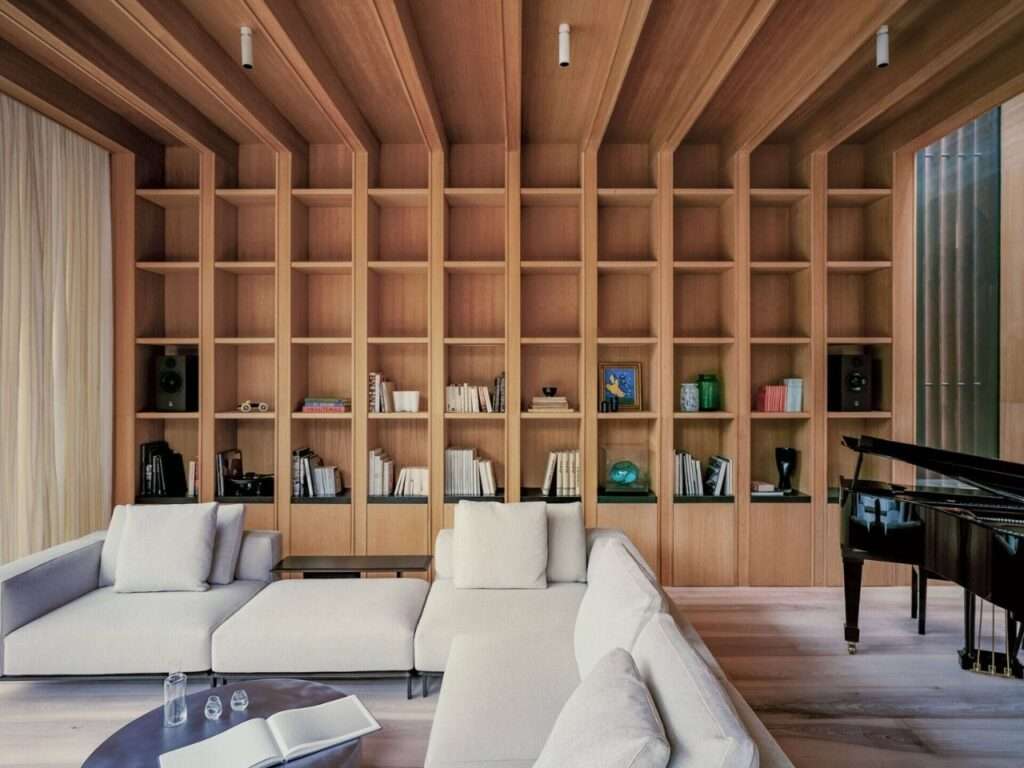 Custom floor-to-ceiling ash wood shelving unit spanning an entire wall behind a modern white sectional sofa and grand piano.
