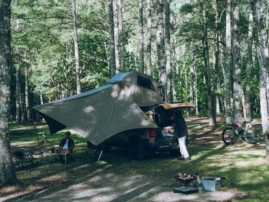 A complete campsite setup in a forest with the Fire Rise RTT and its extended awning used as a social hub.
