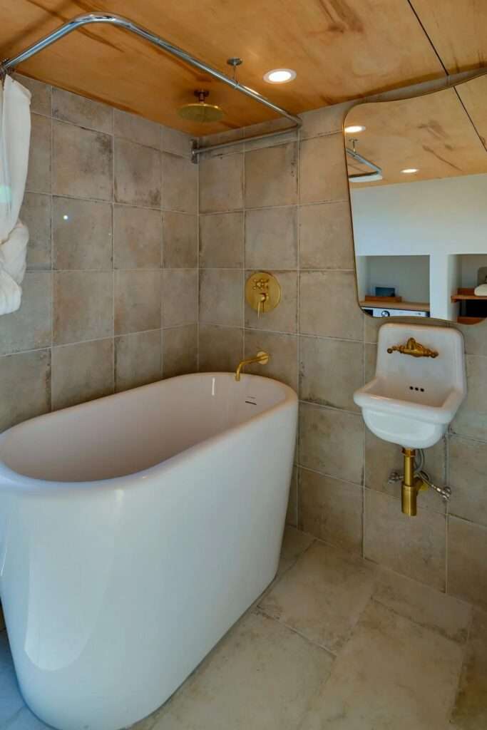 Luxury tiny home bathroom featuring a freestanding white soaking tub, brass fixtures, neutral tiling, and a vintage-style wall-hung sink.