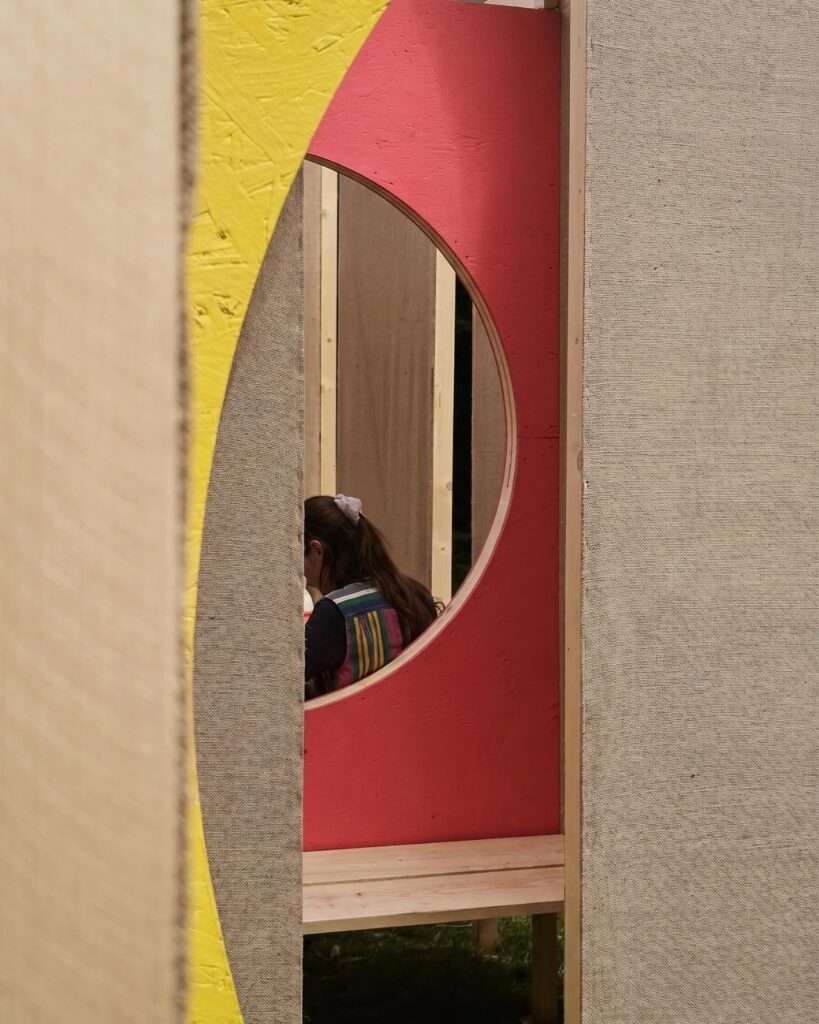 Close-up interior view of the DELULU installation focusing on a circular viewport cutout in a vibrant pink and yellow painted wooden panel, framing a visitor inside the maze.