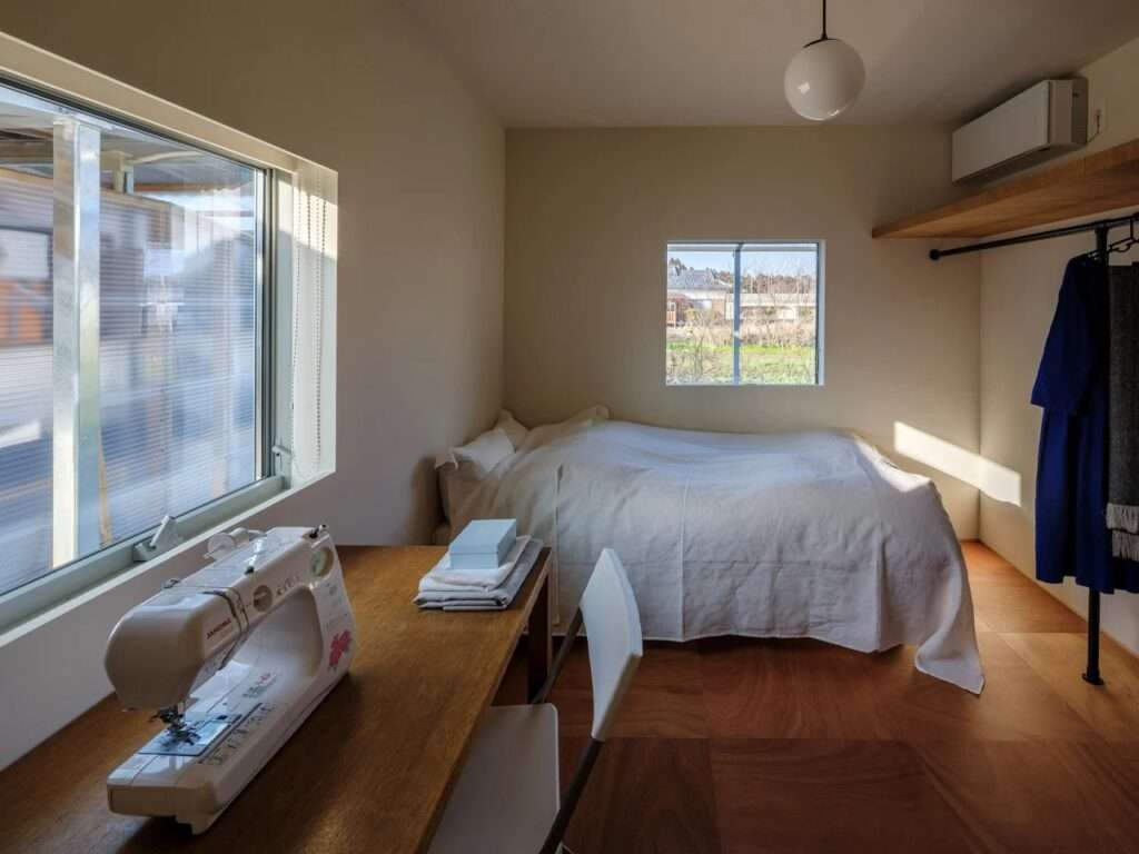 A well-lit contemporary bedroom with a white bed, wooden flooring, a sewing machine on a wooden desk, and two square windows viewing the outdoors.