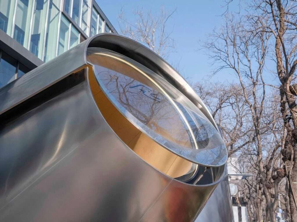 Exterior close-up of the smaller conical cantilevered steel structure with a prominent glass circular window reflecting a modern office building.