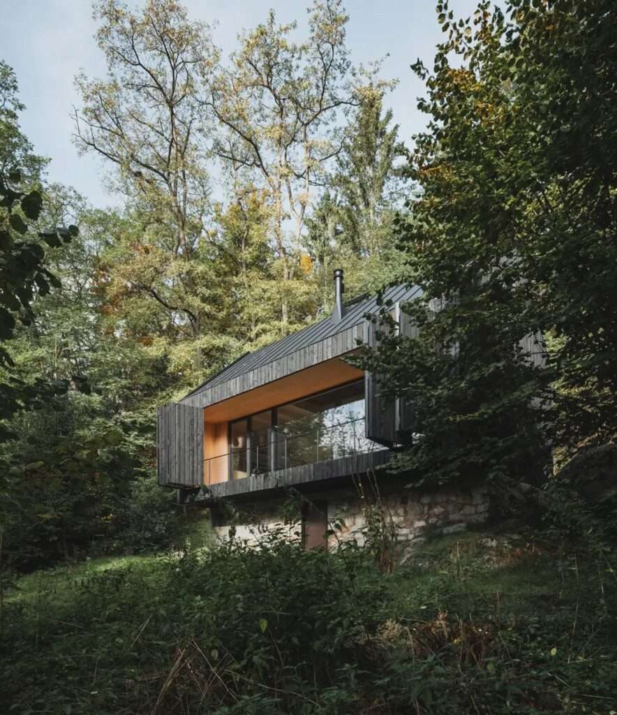 Side elevation of a black wooden cabin showing the contrast between the stone foundation and the large river-facing window.