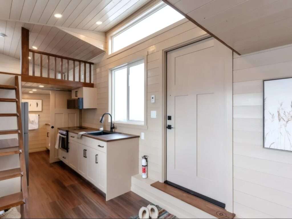 Interior entry view of the Ruby tiny home showing the white front door, a high transom window, and the kitchen counter layout beneath a sloped ceiling.