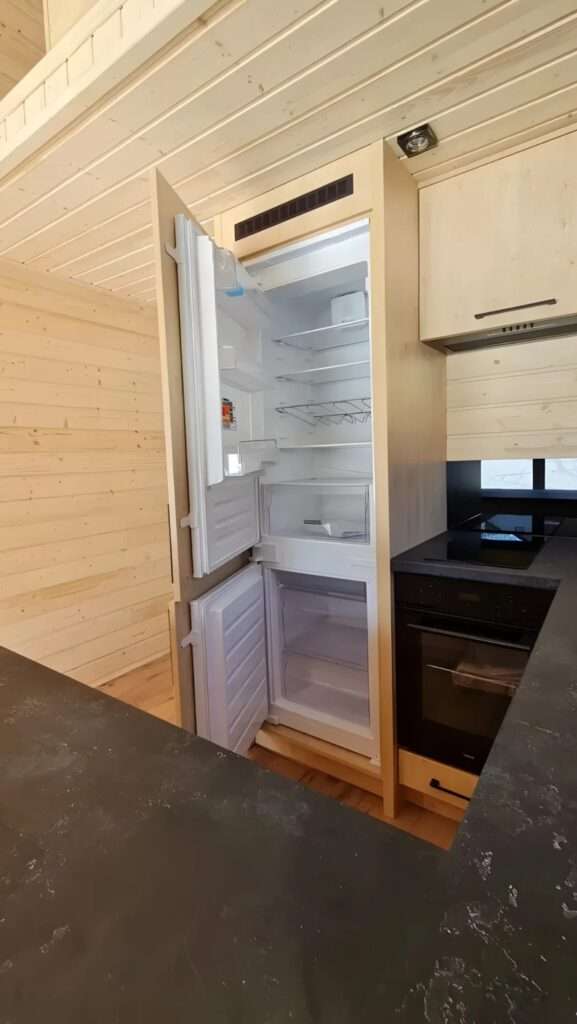 Close-up of a built-in refrigerator and oven within the light wood cabinetry of the Samuel tiny house kitchen.