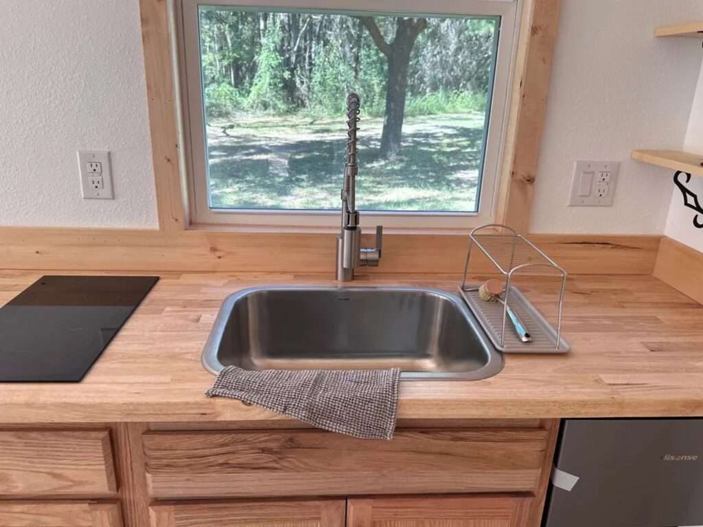 Close-up of a tiny house kitchenette featuring a stainless steel sink, a professional-style faucet, and a large window overlooking trees.