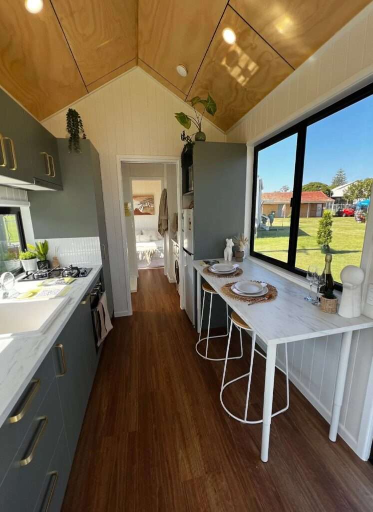 Modern linear kitchen inside Tallebudgera 9.6 featuring dark charcoal cabinetry, white marble-look countertops, gold fixtures, integrated appliances, and a wide horizontal window.