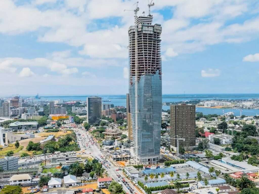 Aerial view of Tour F Tower under construction in Abidjan, detailing the main traffic avenue, the Ebrié Lagoon shoreline, and surrounding mid-century high-rises.