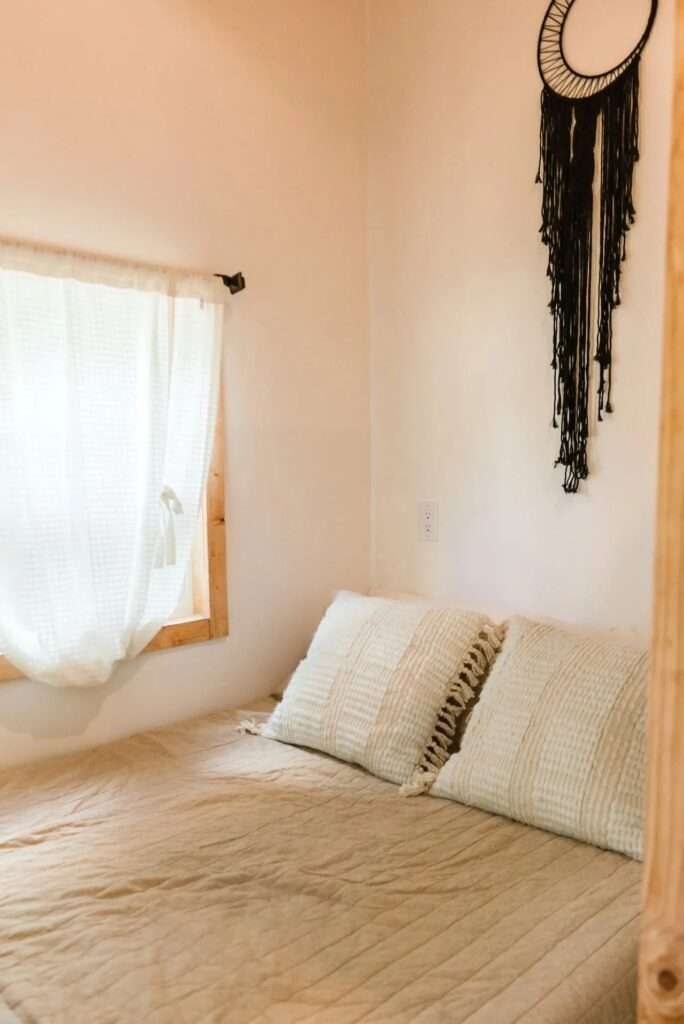 Ground floor bedroom inside the Tulsi tiny home featuring a queen bed under a window with soft white curtains and minimal wall decor.