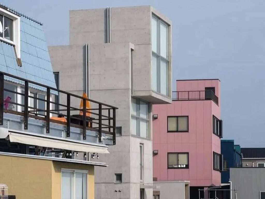 Side profile view of the 4x4 House concrete tower embedded within a dense row of eclectic coastal neighborhood buildings in Kobe.