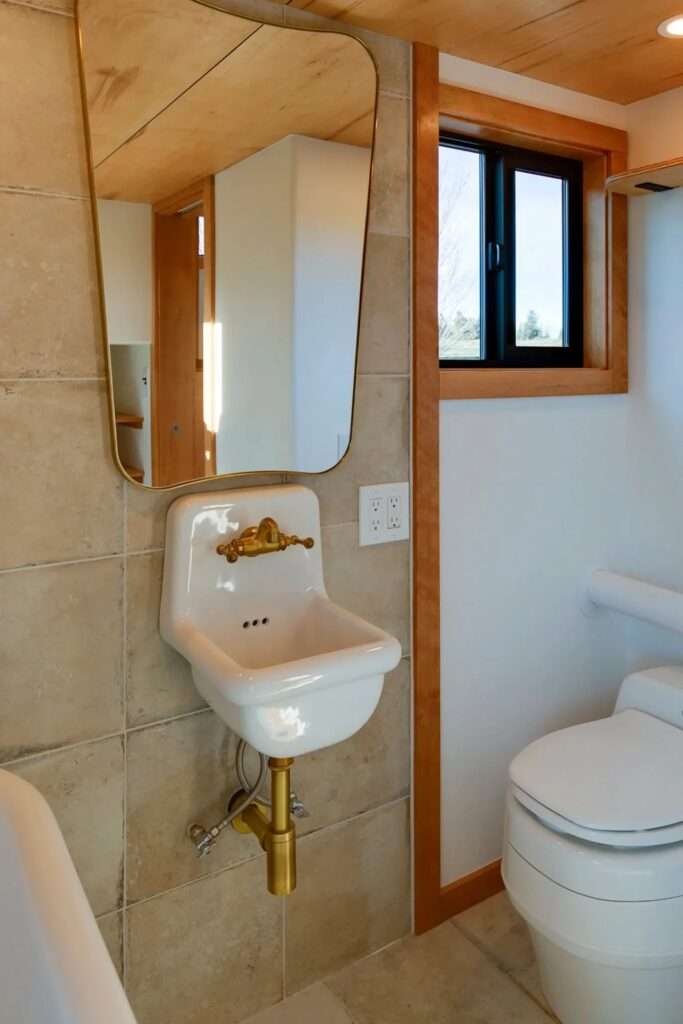 Bathroom interior showing a white porcelain wall-mounted basin with brass plumbing, an asymmetrical mirror, and a modern toilet.