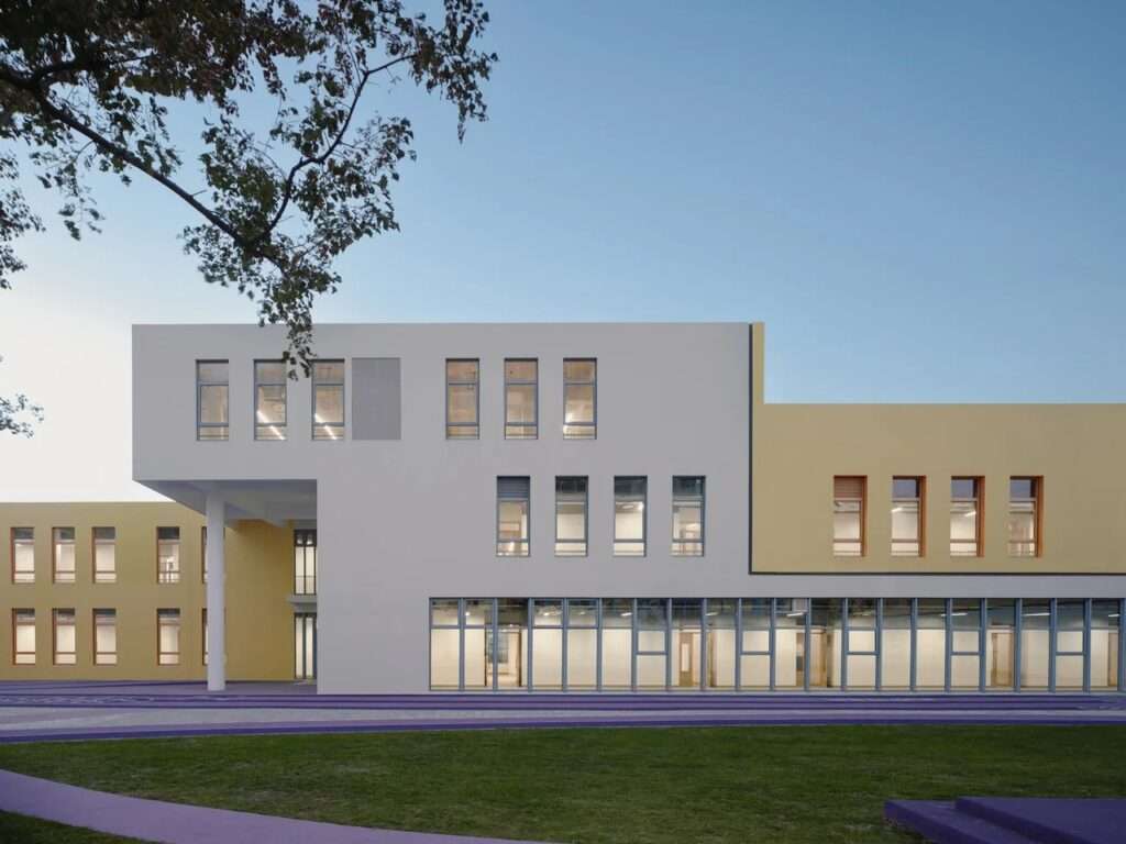 Ground-level view of the modern white and yellow facade of Block Kindergarten featuring ribbon windows, an open pilotis column, and a purple outdoor walkway.