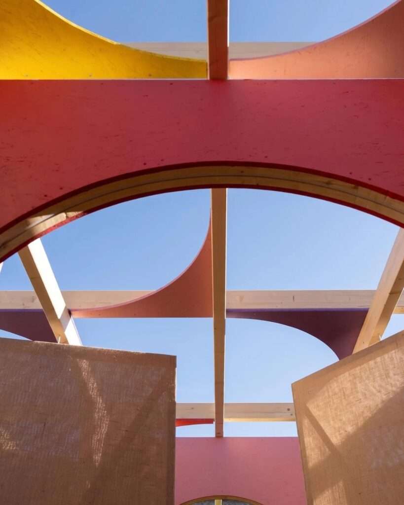 Upward low-angle photograph looking through the open timber ceiling grid of the DELULU maze, featuring curved pink, purple, and yellow accents against a clear blue sky.