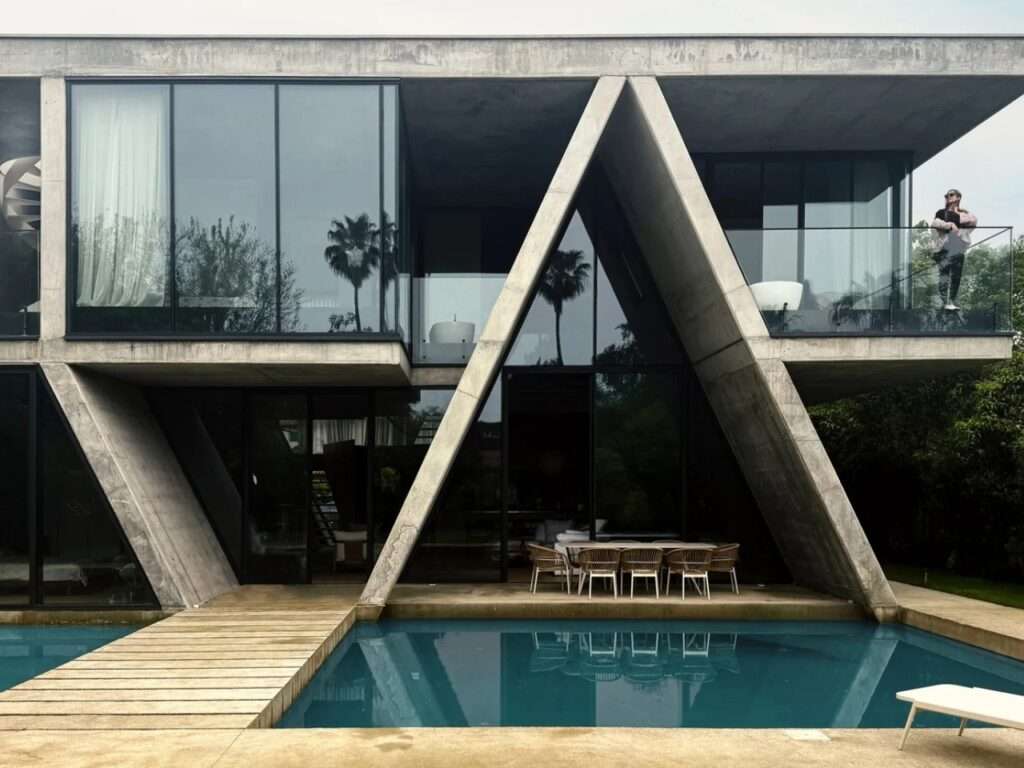 Rear pool-side perspective of Gable Villa highlighting cantilevered upper floors and the central intersecting concrete A-frame canopy under an overcast sky.