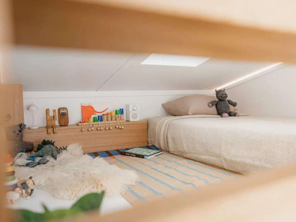 A cozy low-ceiling sleeping loft configured as a child's room with a bed, toys, a skylight, and light wood textures.