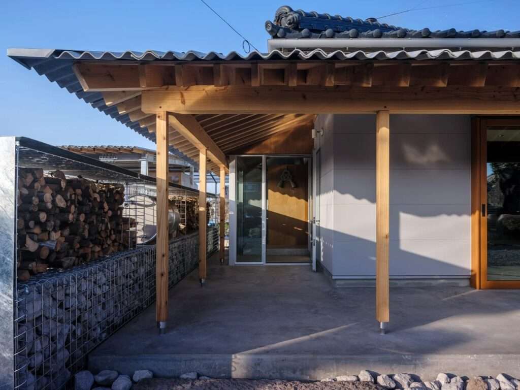 Concrete entrance porch under a heavy timber overhang, adjacent to a stone and wood gabion storage unit against a clear blue sky.