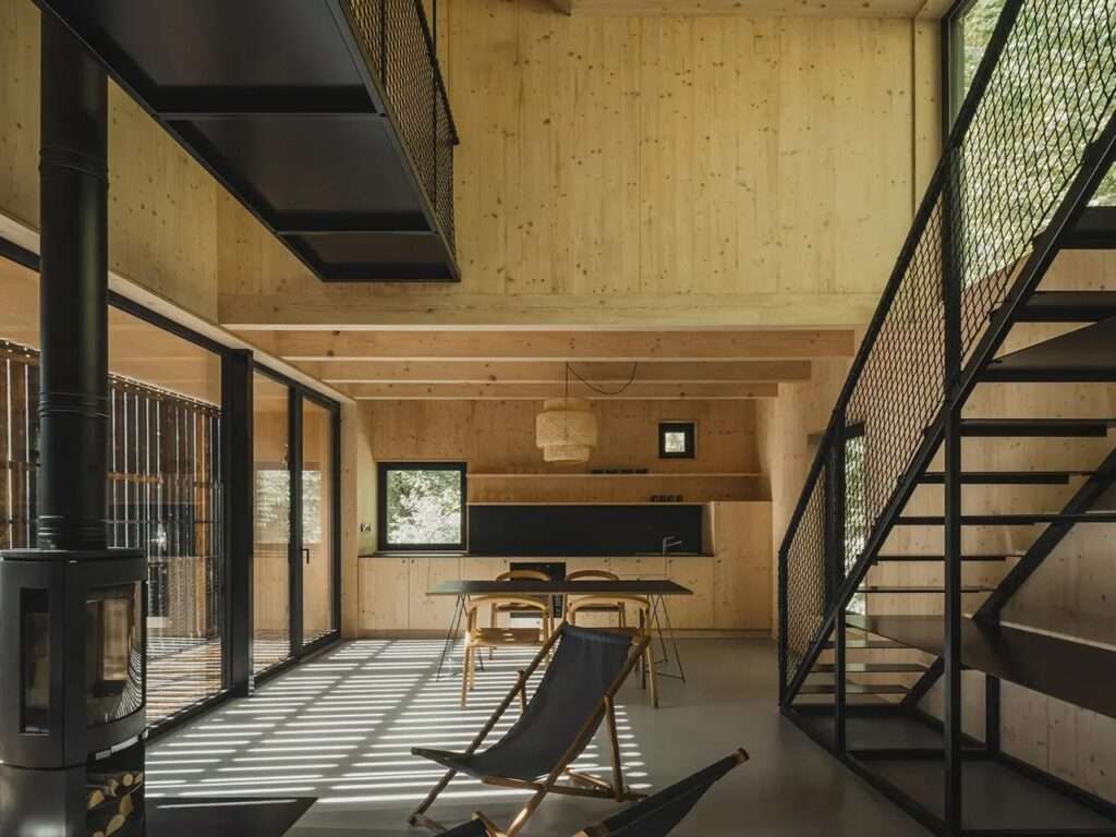 Modern cabin interior featuring light spruce wood walls, a black metal staircase, and sunlight filtering through large glass doors.