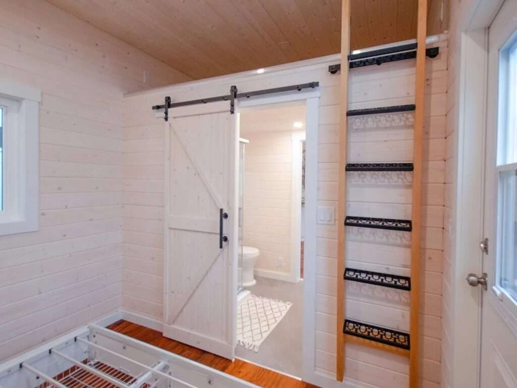 Partially open white rustic sliding barn door revealing a clean white bathroom with a toilet and grey tiled floors in a tiny home.