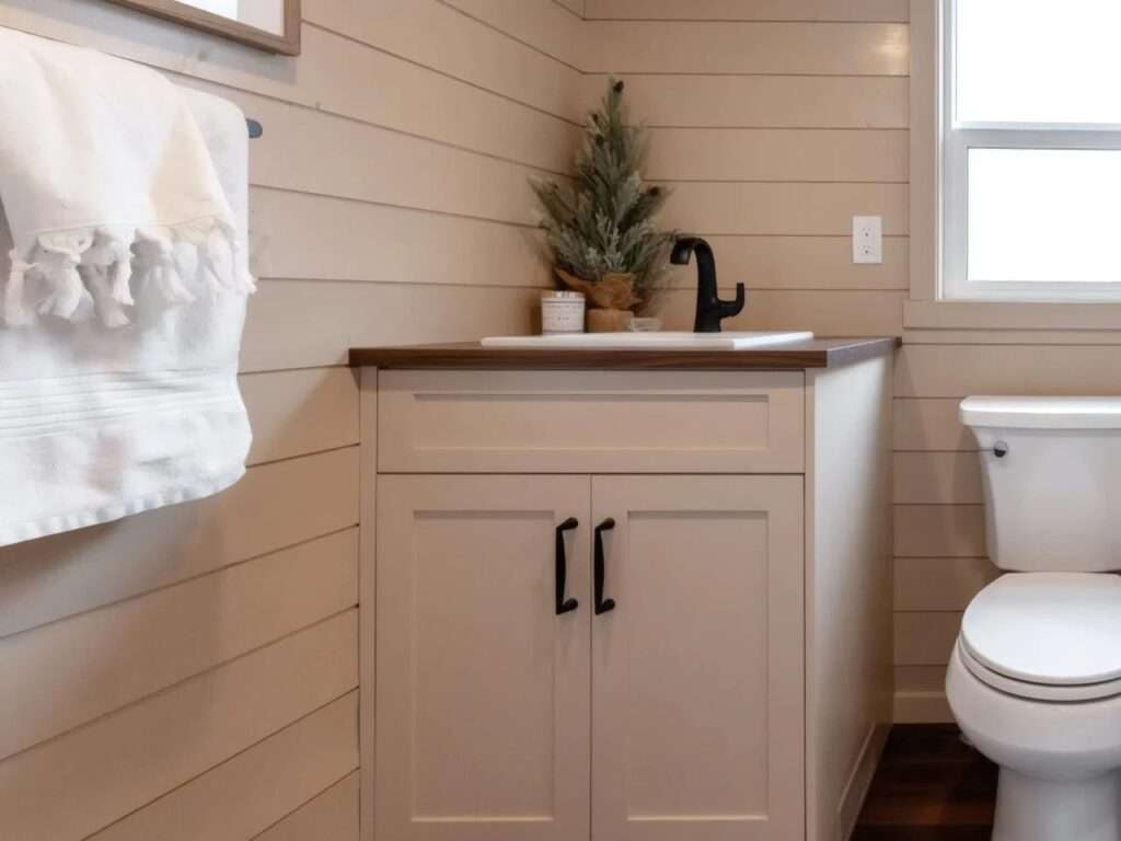 A bathroom inside the Ruby tiny home featuring a white vanity cabinet with a dark wood top, a black faucet, shiplap walls, and a partial view of a white toilet.
