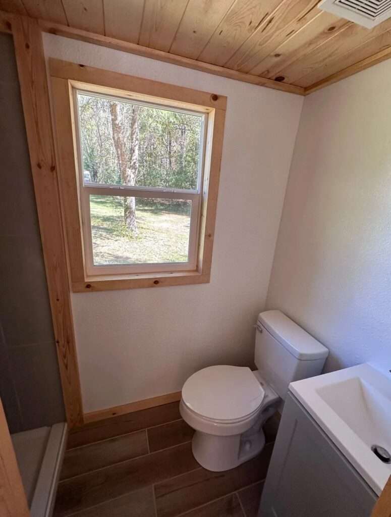 Compact bathroom inside the Shanty tiny house featuring a white toilet, a small vanity, and a window with natural wood framing.