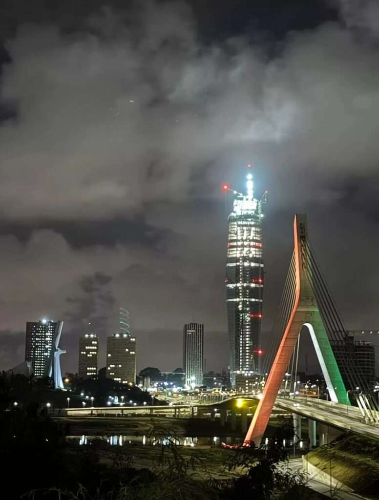 Long exposure night photograph of the Abidjan skyline featuring the brightly lit Tour F Tower structure standing tall next to the illuminated cables of the Cocody bridge.