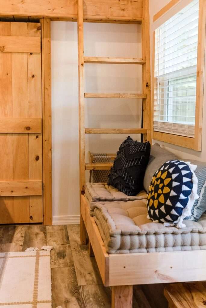 Wooden access ladder standing next to a built-in daybed lounge with textured cushions inside the Tulsi tiny home.