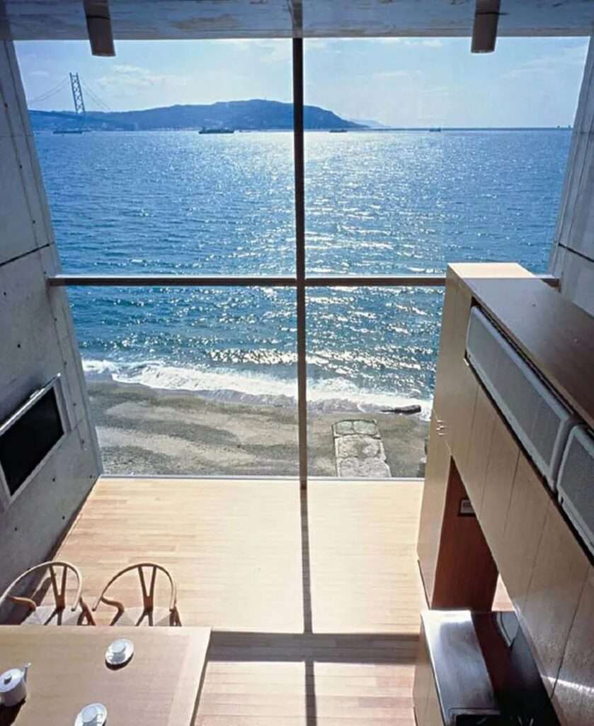 High-angle interior perspective looking through the double-height glass window of the 4x4 House down onto the sandy shoreline and waves below.