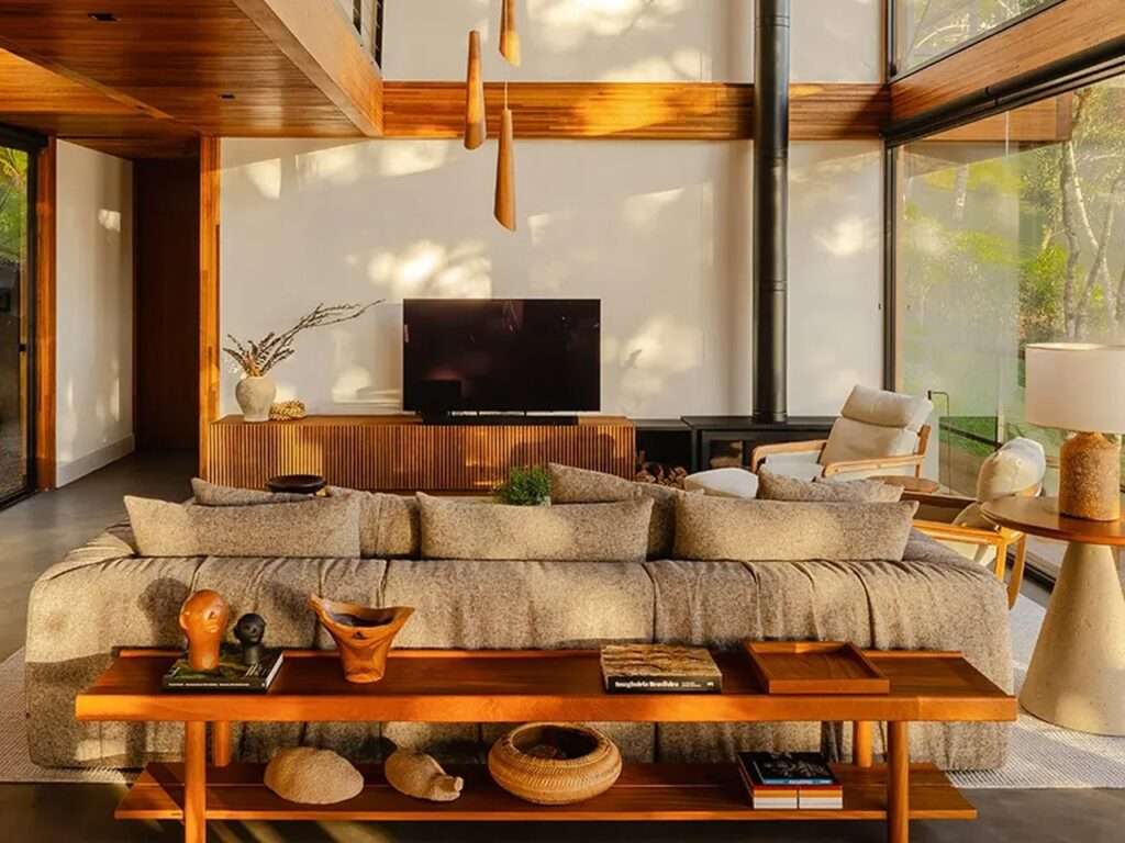 Cozy modern living room interior featuring a grey sofa, wooden furniture, and a black fireplace pipe against a floor-to-ceiling window.