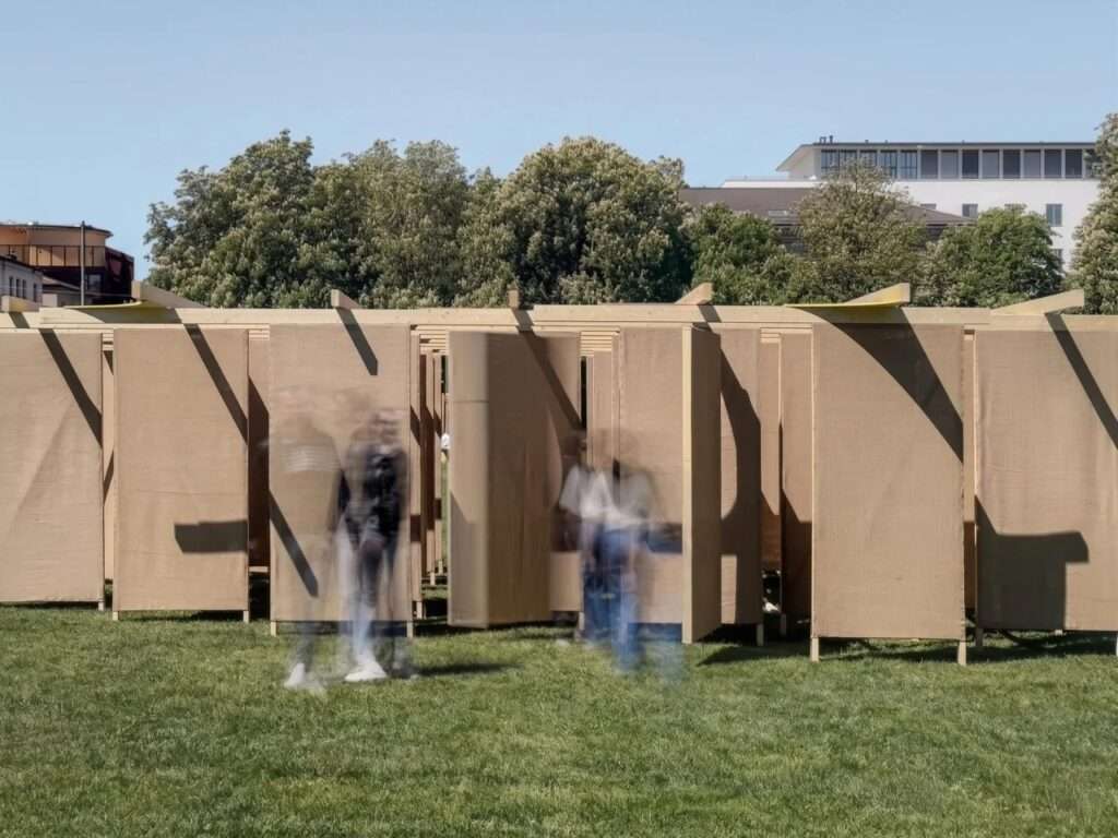 Eye-level photograph with motion blur showing visitors walking through the shifting rows of vertical jute fabric screens of the DELULU installation on a sunny day.