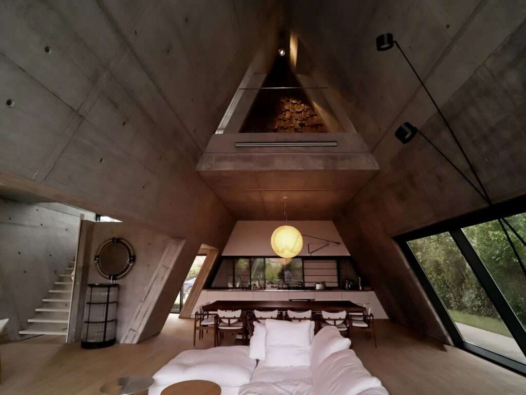 Wide-angle interior shot of a luxury minimalist living room with a white sofa under a dramatic, sloping raw concrete ceiling.