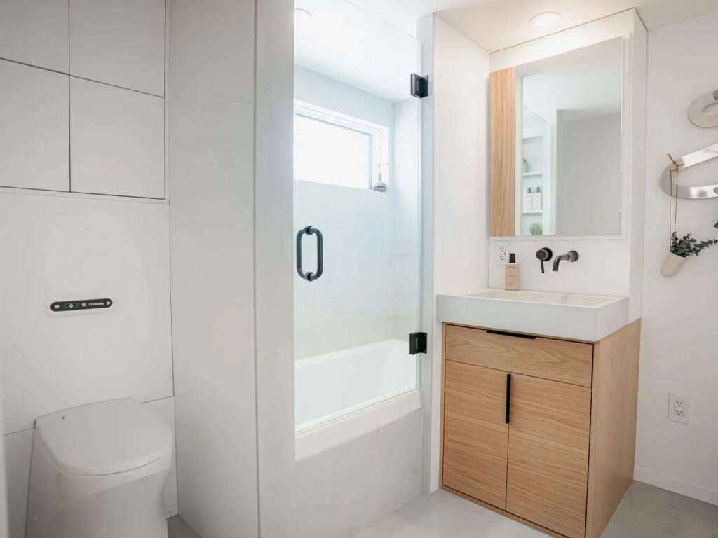 A modern white bathroom inside a tiny home featuring a wood vanity, black fixtures, a toilet, and a glass-enclosed deep soaking bathtub.