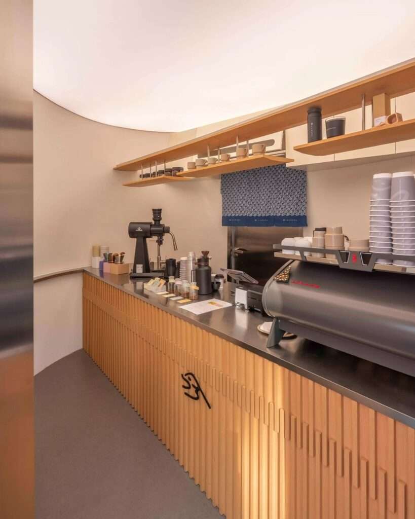 Internal perspective of the Kurasu coffee bar showing the counter setup, professional coffee grinder, and the minimalist Kurasu logo on the wooden slats.