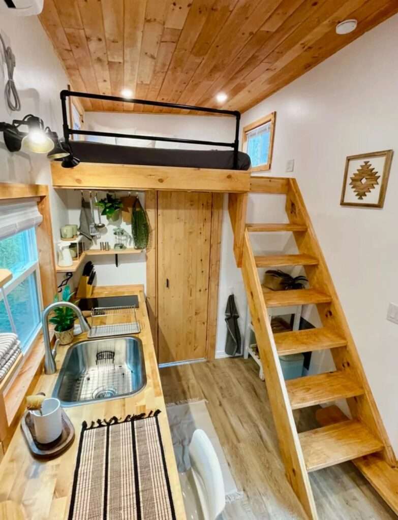 Top-down view from the sleeping loft into the kitchen and entry area of Rasa tiny home, showcasing wooden flooring and stairs.