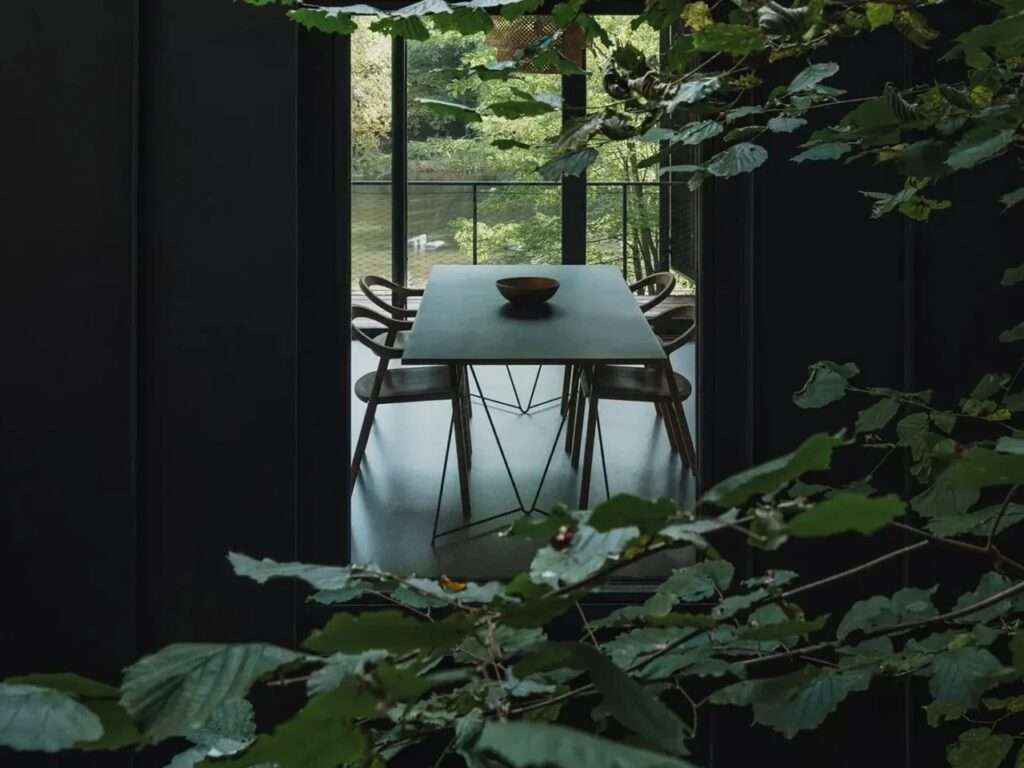 View through green leaves looking into a minimalist dining room with a black table and chairs overlooking the river.