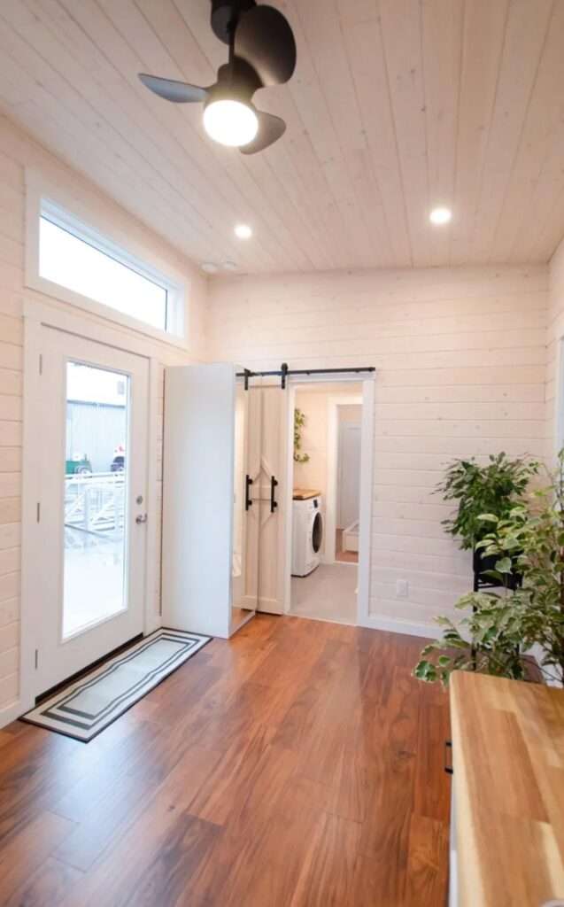Open-concept tiny home living room with rich walnut wood flooring, white glass entry door, high ceilings, and a modern black ceiling fan.