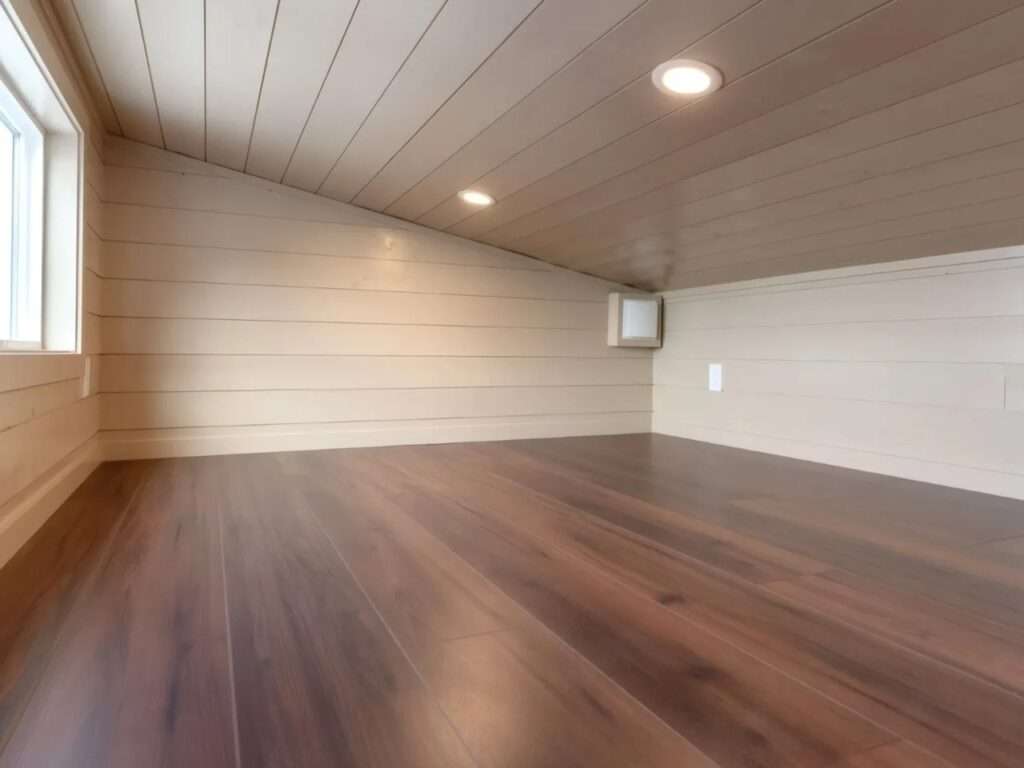 Empty loft bedroom inside the Ruby tiny home featuring hardwood flooring, sloped shiplap ceilings with recessed LED lighting, and a side window.