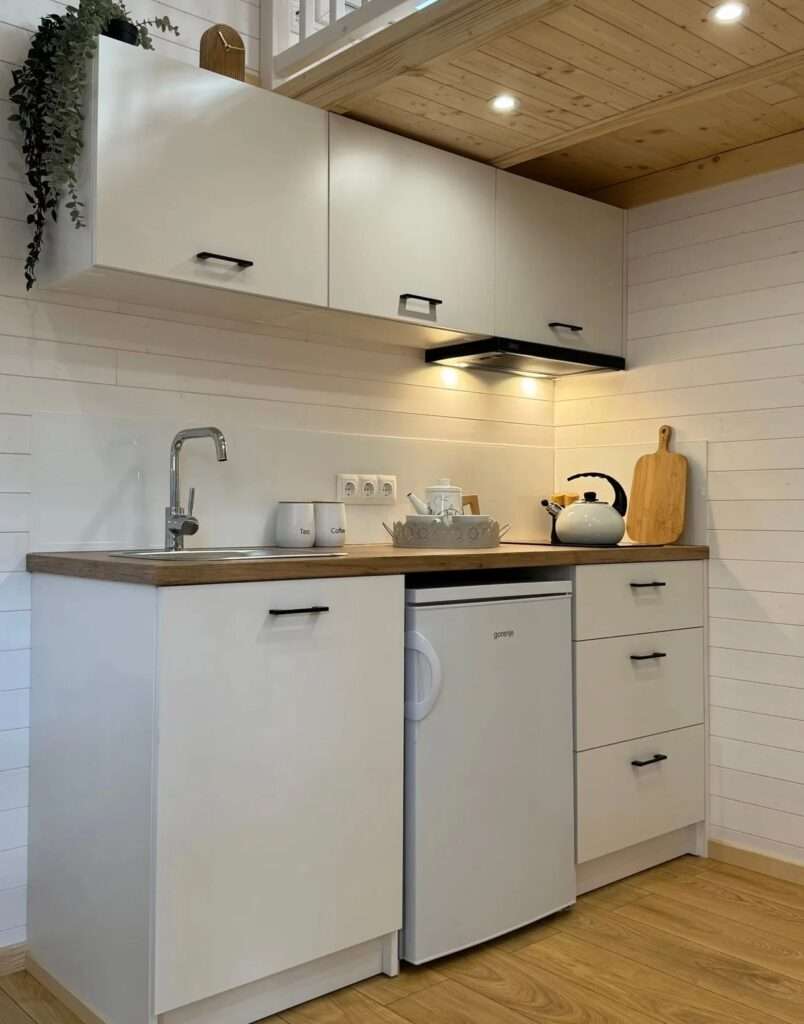 Modern white kitchenette in a tiny house featuring a wooden countertop, small fridge, and integrated sink.
