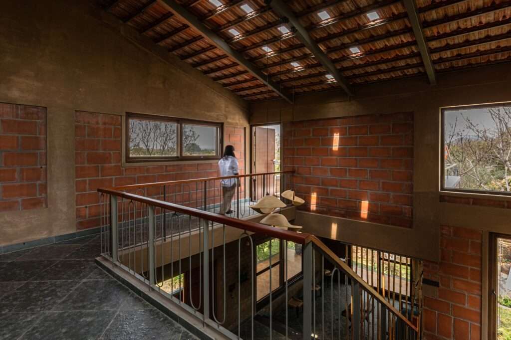 Architectural interior showing a person walking across an upper level metal bridge walkway with open railings over a double height courtyard with mud brick walls.
