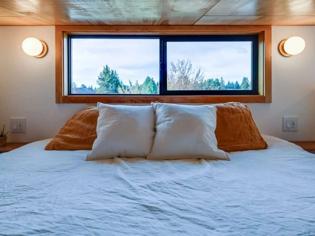 Cozy loft bedroom inside a tiny house featuring a large bed with white linens, warm spherical wall sconces, and a horizontal awning window.