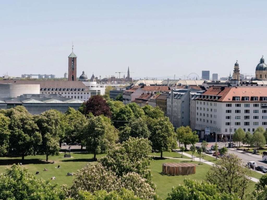 High-angle distant view of the Munich urban cityscape, showing the DELULU installation as a small timber pavilion on a large green park lawn surrounded by trees and historic city towers.