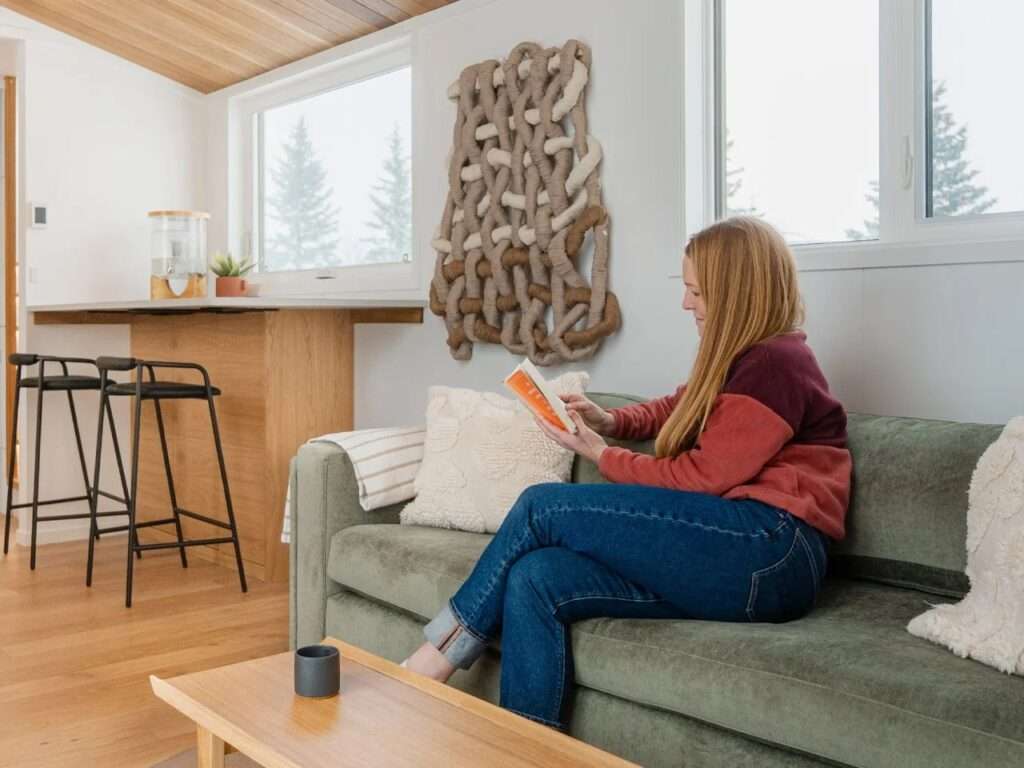 A woman sitting on an olive green sofa reading a book next to large windows and a custom woven wall hanging in the Halcyon Grand.
