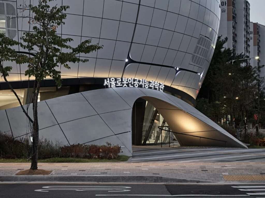 Close-up dusk view of the main entrance arch of RAIM with glowing signage in Korean characters beneath the curved metallic panel facade.